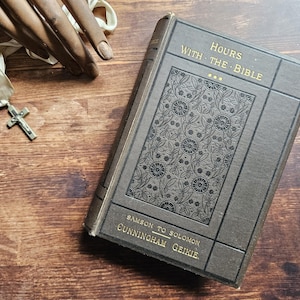Könnte beinhalten: Ein antikes Buch mit dem Titel "Hours with the Bible" mit goldener Schrift und einem dekorativen floralen Muster auf dem Einband. Ein silbernes Kreuz an einem weißen Band liegt neben dem Buch auf einer Holzoberfläche.