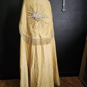 May include: A long, golden-yellow religious vestment with intricate embroidery. The back features a detailed design of a dove within a sunburst, with gold fringe along the bottom. The garment is displayed on a mannequin.