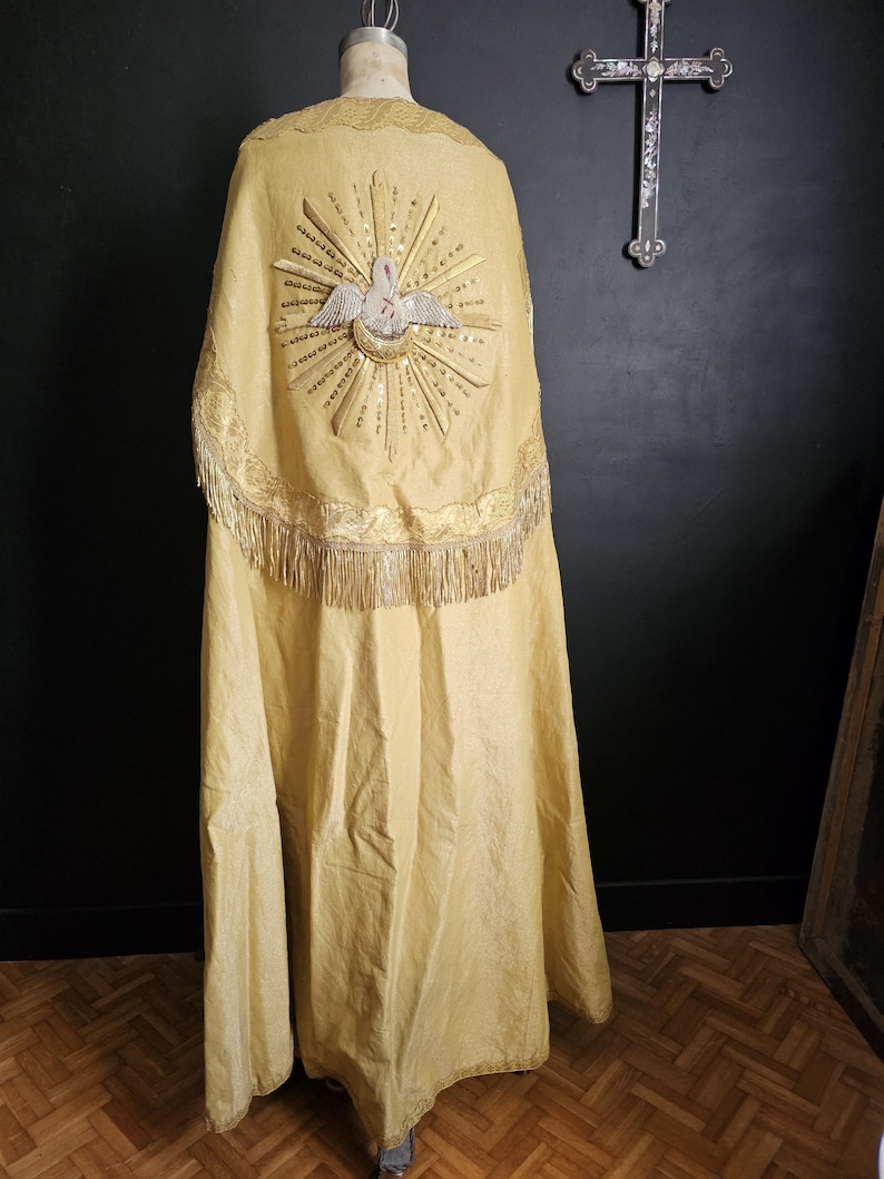 May include: A long, golden-yellow religious vestment with intricate embroidery. The back features a detailed design of a dove within a sunburst, with gold fringe along the bottom. The garment is displayed on a mannequin.
