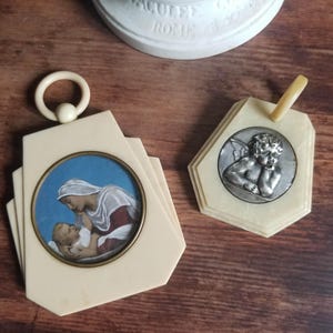Op de afbeelding: Twee vintage ivoor-kleurige religieuze medaillons. Eén heeft een zilverkleurig beeld van een cherubijn met vleugels. De andere heeft een geschilderd beeld van de Maagd Maria die een baby Jezus vasthoudt.