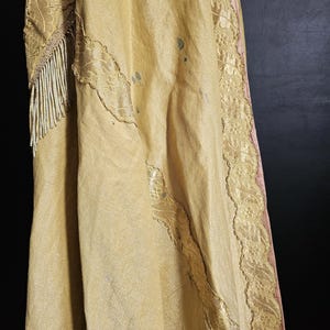 May include: Close-up of a vintage, gold-colored garment with intricate lace detailing and beaded fringe. The fabric has a subtle sheen and appears to be aged. The edges are trimmed with delicate lace, and the garment has some visible stains.