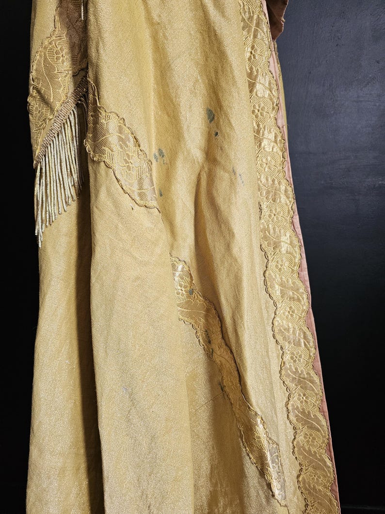 May include: Close-up of a vintage, gold-colored garment with intricate lace detailing and beaded fringe. The fabric has a subtle sheen and appears to be aged. The edges are trimmed with delicate lace, and the garment has some visible stains.