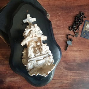 Può includere: Un'antica decorazione murale religiosa con una scena della natività scolpita, una croce e un supporto in legno nero. Un rosario, una piccola carta e una chiave sono presenti anche su una superficie di legno.