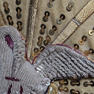 May include: Close-up of a detailed textile with gold, silver, and purple embroidery. The design features a stylized bird or angel with intricate wing and body details. Gold sequins and metallic threads add texture and shine to the fabric.