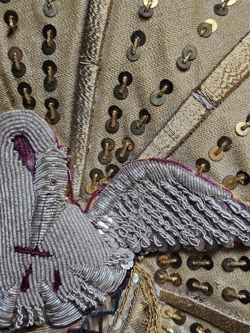 May include: Close-up of a detailed textile with gold, silver, and purple embroidery. The design features a stylized bird or angel with intricate wing and body details. Gold sequins and metallic threads add texture and shine to the fabric.