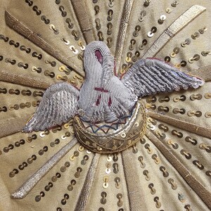 May include: Close-up of an embroidered design featuring a dove with outstretched wings, set against a gold fabric background. The dove is white with silver wings and a red accent. Gold sequins and radiating gold bands add detail.