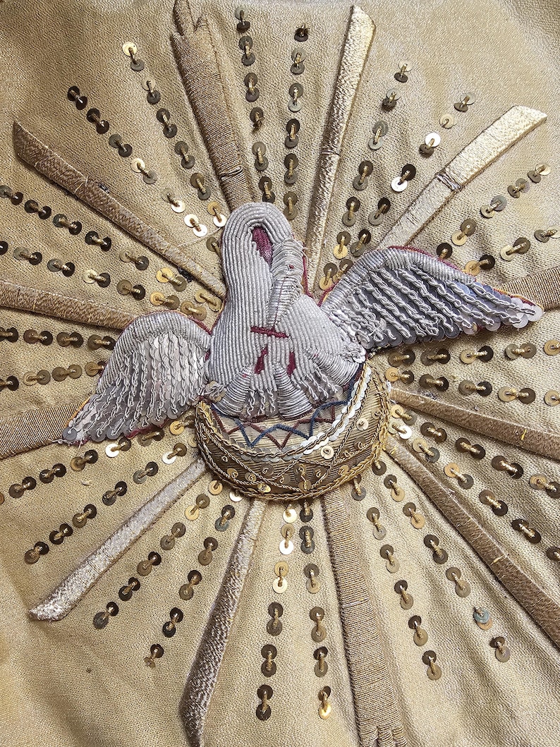 May include: Close-up of an embroidered design featuring a dove with outstretched wings, set against a gold fabric background. The dove is white with silver wings and a red accent. Gold sequins and radiating gold bands add detail.