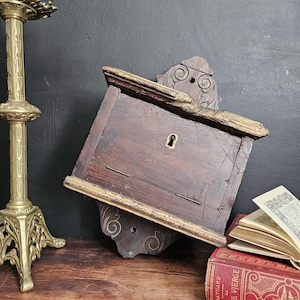 May include: A dark wooden antique wall-mounted box with a keyhole, ornate carvings, and gold trim. A brass candlestick and a red book with gold lettering are also visible.