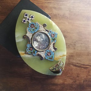 Peut inclure: Un objet décoratif de forme ovale vert avec un motif en argent et émail représentant un motif floral et un camée d'un visage de femme. L'objet a une petite poignée ornée de couleur dorée.