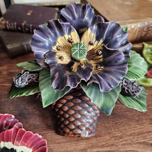 Op de afbeelding: Een decoratief keramisch object met een grote, gegolfde paars-gouden bloem met een groene zaaddoos in het midden. De bloem is omringd door groene bladeren en donkere knoppen, rustend op een bruine, getextureerde basis met een dennenappelpatroon.