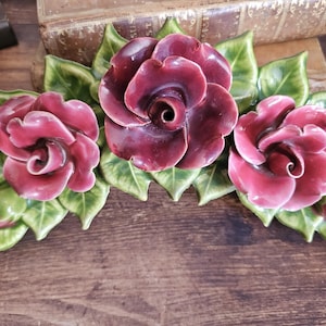 May include: A decorative ceramic wall plaque featuring three sculpted deep burgundy roses surrounded by glossy green leaves. The piece has a curved, horizontal arrangement with detailed petals and foliage, finished with a smooth, reflective glaze.