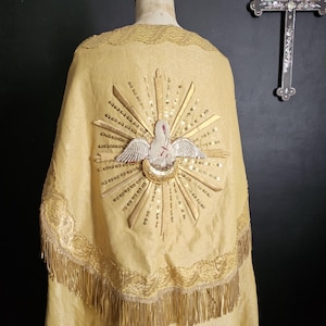 May include: A gold-colored religious vestment with a detailed embroidered design. The back features a dove with a halo, surrounded by rays and sequins. The trim includes lace and fringe. A cross hangs in the background.