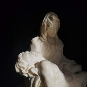 May include: A white statue of a woman holding a man in her arms. The woman is wearing a long robe and has her head bowed. The man is lying on her lap with his head resting on her shoulder.