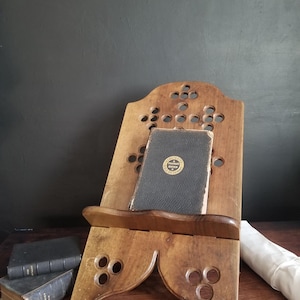 May include: A wooden book stand with a dark finish, holding a vintage book. The stand has decorative cut-out circles and a shelf. Two other books are visible on the wooden surface. The book on the stand has a gold emblem.