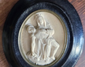 May include: A circular, black framed relief sculpture showing a religious scene. The sculpture features two figures in a cream colour, set against a cream background. The frame is a dark, matte black.