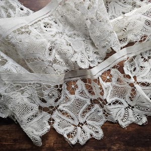 May include: Close-up of white lace fabric with intricate floral patterns. The lace is folded and layered, creating depth and texture. The edges have a scalloped design. The fabric is likely used for crafting, sewing, or home decor.