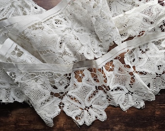 May include: Close-up of white lace fabric with intricate floral patterns. The lace is folded and layered, creating depth and texture. The edges have a scalloped design. The fabric is likely used for crafting, sewing, or home decor.