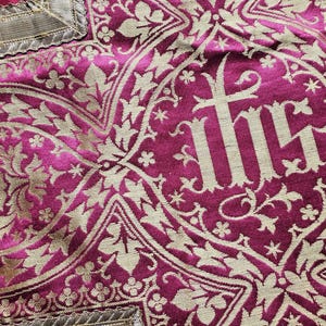 May include: Close-up of a magenta textile with a gold floral pattern. The design includes the letters IHS in a central medallion, surrounded by ornate leaves and flowers. The fabric has a textured appearance.