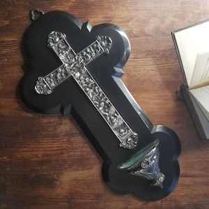 Op de afbeelding: Een zwart wandkruis met een zilveren kruisoverlay, met gedetailleerde reliëfsnijwerken. Het kruis heeft een decoratieve basis en wordt weergegeven op een houten oppervlak. Op de achtergrond is een open boek te zien.
