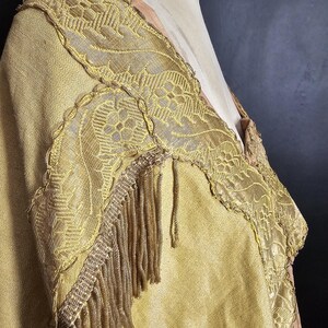 May include: A vintage gold-colored garment with intricate lace detailing and fringe trim. The fabric appears to be a textured weave, and the garment is displayed on a mannequin. The design features floral patterns and a layered construction.