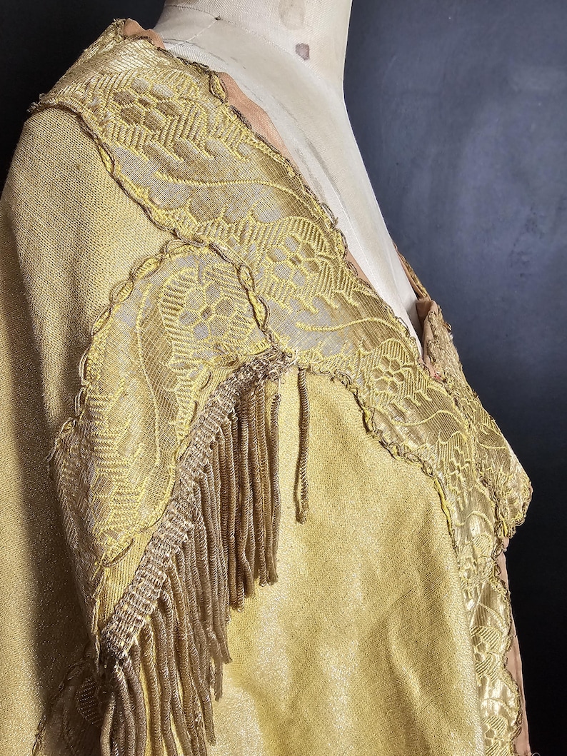 May include: A vintage gold-colored garment with intricate lace detailing and fringe trim. The fabric appears to be a textured weave, and the garment is displayed on a mannequin. The design features floral patterns and a layered construction.
