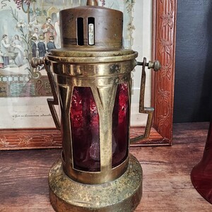 May include: Vintage brass lantern with a cross finial. The lantern has a red glass panel and a handle. The lantern is on a wooden surface. The lantern is an example of religious decor.