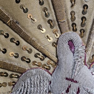 May include: Close-up of a textile with a detailed embroidered design. The fabric is a muted gold color, adorned with gold sequins and intricate silver and purple embroidery, possibly a decorative element.