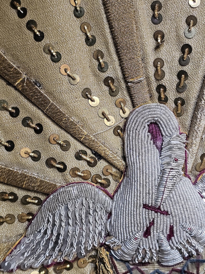 May include: Close-up of a textile with a detailed embroidered design. The fabric is a muted gold color, adorned with gold sequins and intricate silver and purple embroidery, possibly a decorative element.