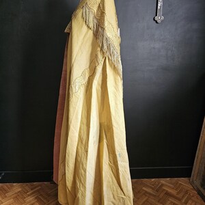 May include: A vintage, golden-yellow cape with intricate embroidery and fringe detailing. The cape is draped over a mannequin, showcasing its flowing design and texture. The garment appears to be made of a lightweight fabric, with a contrasting pink lining visible.