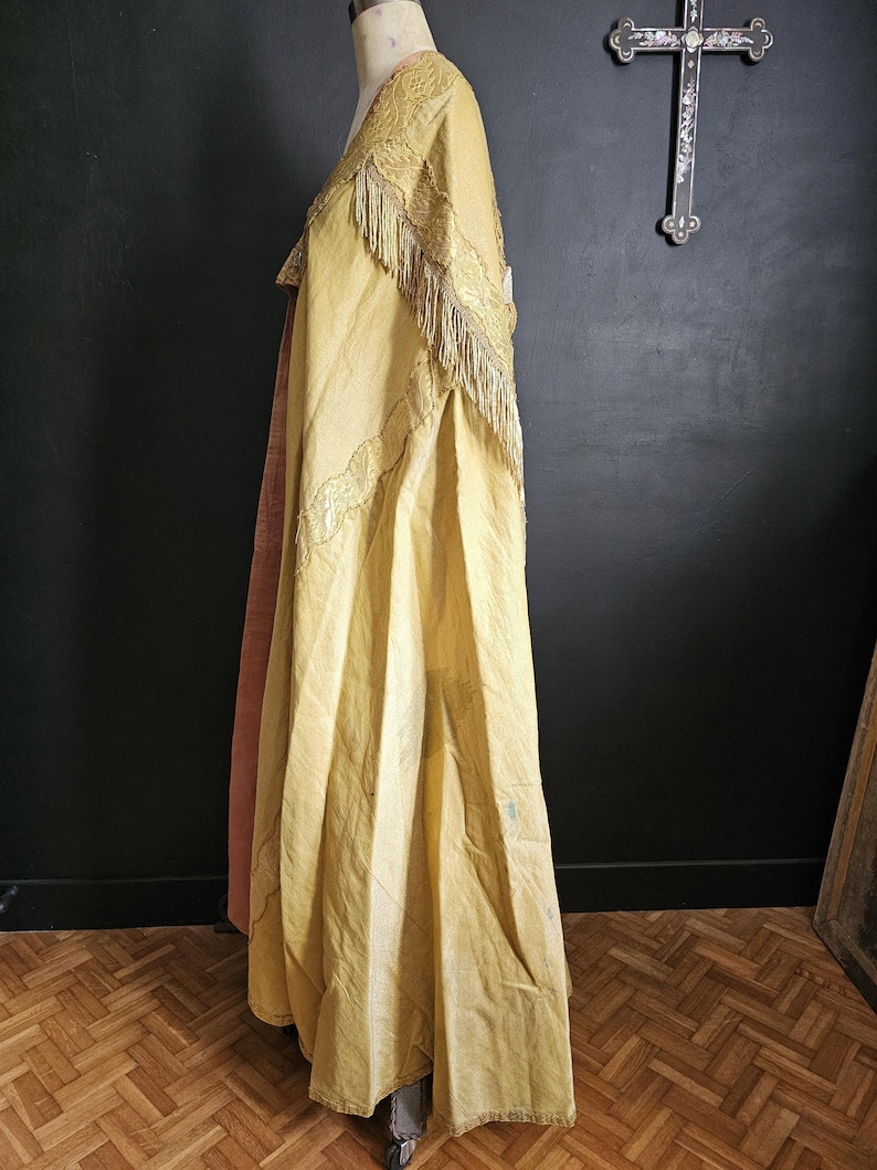 May include: A vintage, golden-yellow cape with intricate embroidery and fringe detailing. The cape is draped over a mannequin, showcasing its flowing design and texture. The garment appears to be made of a lightweight fabric, with a contrasting pink lining visible.