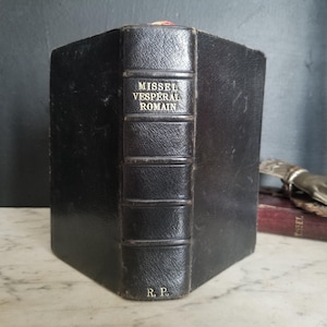 May include: A black leather-bound book with gold lettering on the spine that reads "MISSEL VESPERAL ROMAIN." The book is closed and sitting on a white marble surface.