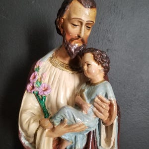 May include: A vintage statue of Saint Joseph holding a small child. The statue is made of painted plaster and features a realistic depiction of the figures. Saint Joseph is wearing a brown robe and a white tunic, while the child is wearing a blue tunic. The statue is in good condition with some minor wear and tear.