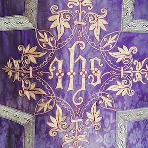 May include: Close-up of a purple liturgical vestment with gold and red embroidery. The design features floral patterns and the letters IHS in the center. Silver trim borders the edges of the vestment.
