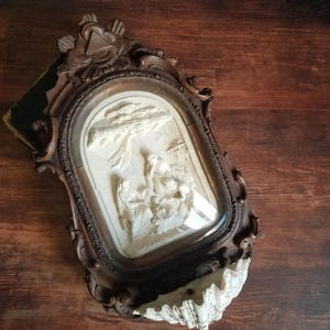 Può includere: Un'antica scultura a rilievo, ornata, con cornice marrone scuro. Il rilievo raffigura una scena religiosa in bianco, su uno sfondo color crema. La cornice presenta intagli intricati e una parte superiore decorativa. Il pezzo è posto su una superficie di legno.