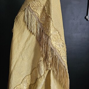 May include: A gold-colored shawl with intricate lace trim and a fringe border. The fabric has a subtle sheen, and the lace features a detailed floral pattern. The shawl is draped, showcasing its texture and design.