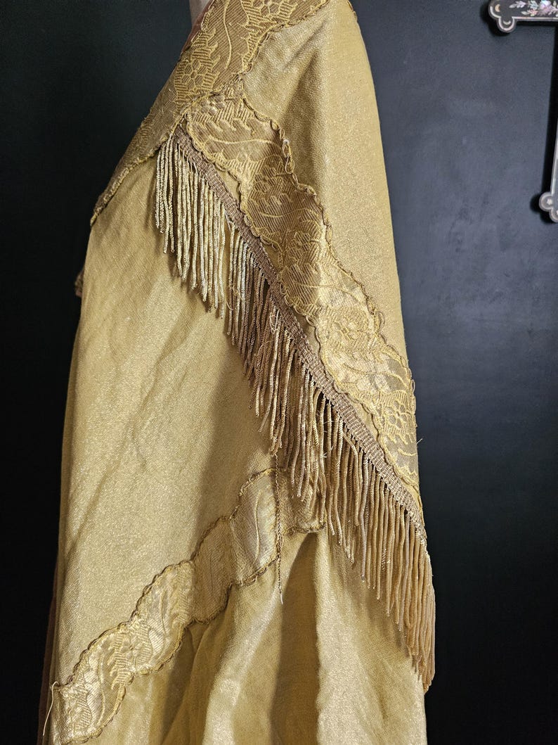 May include: A gold-colored shawl with intricate lace trim and a fringe border. The fabric has a subtle sheen, and the lace features a detailed floral pattern. The shawl is draped, showcasing its texture and design.