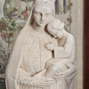 May include: A matte off-white stone sculpture depicting a crowned figure holding a small child. The figure wears a long, draped garment with a veil, while the child rests against the figure's shoulder. The surface has a textured, weathered appearance.