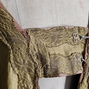 May include: Close-up of a vintage garment featuring intricate gold-colored embroidery and trim. The design includes floral patterns and decorative metal clasps. The garment appears to be made of a shimmering, light-colored fabric with a pink lining.