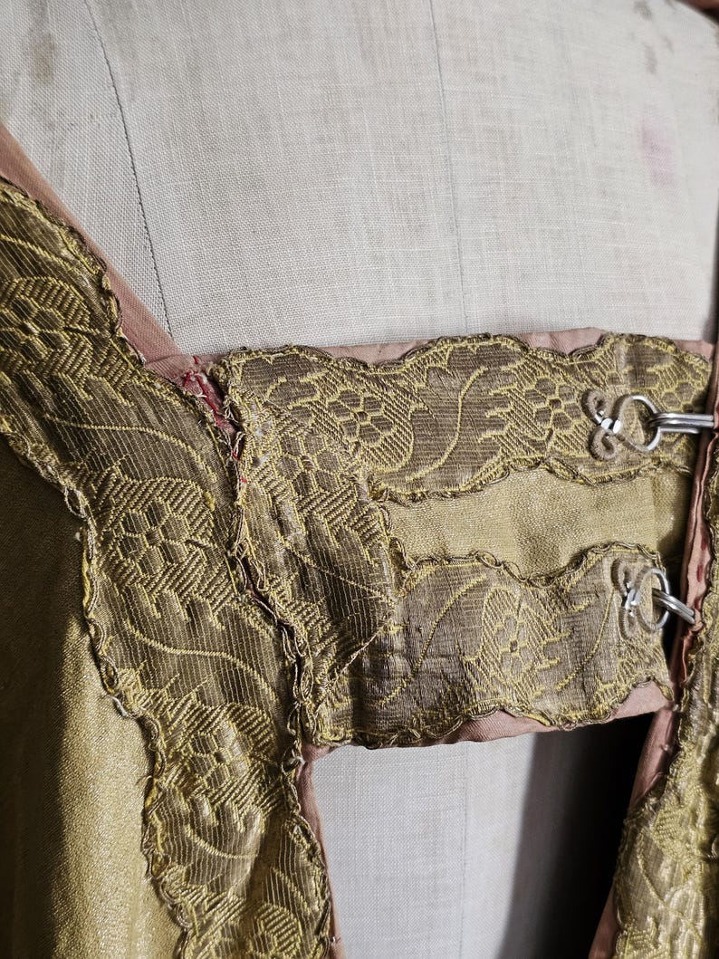 May include: Close-up of a vintage garment featuring intricate gold-colored embroidery and trim. The design includes floral patterns and decorative metal clasps. The garment appears to be made of a shimmering, light-colored fabric with a pink lining.