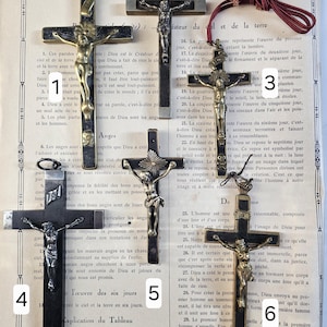 May include: Collection of antique crucifixes. Each cross features a detailed depiction of Jesus Christ. The crosses are made of dark metal and gold-toned metal, with varying designs and sizes. The background is a vintage book page with French text.
