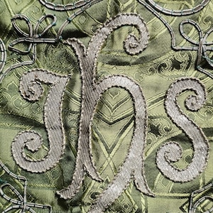 May include: Close-up of a green fabric with a repeating pattern and ornate silver embroidery. The embroidery forms the letters IHS, with decorative swirls and flourishes. The fabric has a subtle sheen.