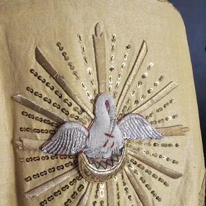 May include: A gold-colored religious garment with an embroidered dove emblem. The dove is white with silver wings and a red cross, surrounded by gold rays and sequins. The garment has a decorative gold trim.