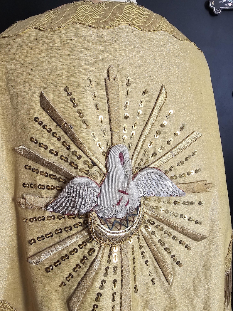 May include: A gold-colored religious garment with an embroidered dove emblem. The dove is white with silver wings and a red cross, surrounded by gold rays and sequins. The garment has a decorative gold trim.