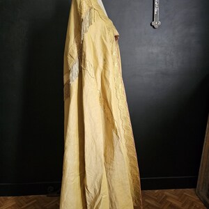 May include: A long, flowing, golden-yellow cape with intricate embroidery and fringe detailing. The cape is draped over a mannequin, showcasing its elegant design and texture. The garment appears to be vintage or antique, with a focus on craftsmanship.