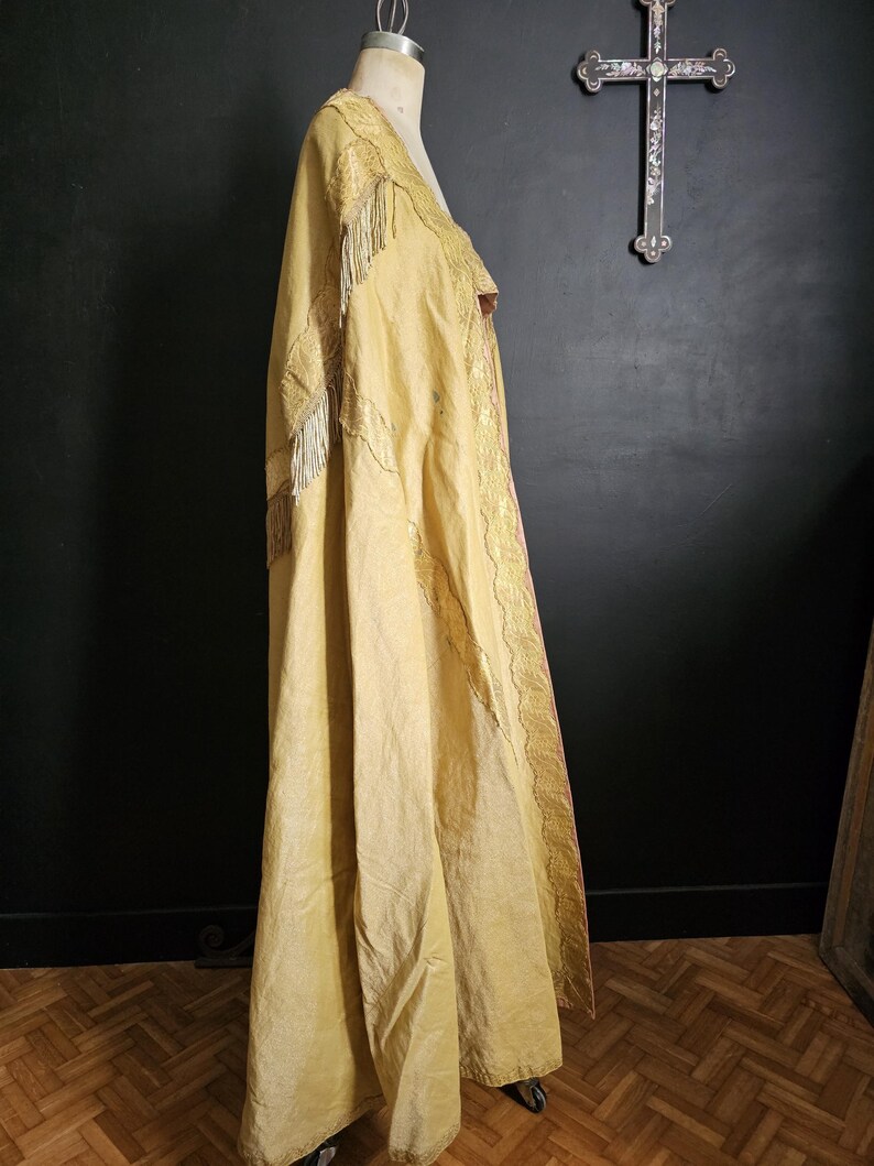 May include: A long, flowing, golden-yellow cape with intricate embroidery and fringe detailing. The cape is draped over a mannequin, showcasing its elegant design and texture. The garment appears to be vintage or antique, with a focus on craftsmanship.