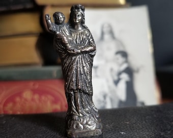 May include: A silver-toned metal figurine of the Virgin Mary holding a baby Jesus. The figurine is standing on a black surface.