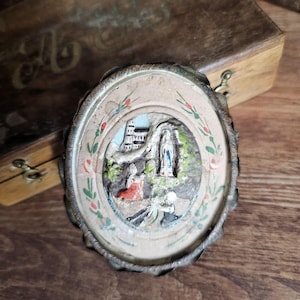 May include: A vintage, circular religious medallion featuring a detailed relief scene. The central image shows a grotto with figures, painted in muted tones. The medallion has a decorative, scalloped metal edge and floral accents.