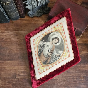 May include: Antique religious artwork featuring a diamond-shaped image of a saint holding lilies, set within a red velvet frame. The artwork is surrounded by a gold border and rests on a wooden surface, with antique books and a metal sculpture in the background.