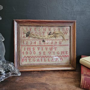 以下が含まれることがあります： ベージュ色の生地に赤いステッチが施された、額装されたアンティークのクロスステッチのサンプル。 サンプルには文字、数字、テキスト「INSTITUT 1930 SEVIGNE M.CAILLARD」が含まれています。 三つ編みの髪の毛が取り付けられています。 ガラスの燭台と本が背景にあります。