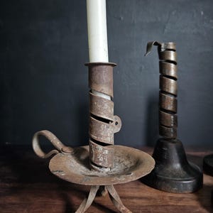 Peut inclure: Deux bougeoirs en métal avec une finition rouillée. Le bougeoir au premier plan présente un design en spirale, une coupelle circulaire, une petite poignée latérale et trois pieds. Une bougie blanche est insérée dans le tube en métal.